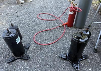 Two HVAC compressors on an asphalt driveway next to a red pressurized gas cylinder.