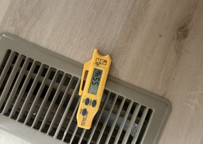 A yellow digital thermometer resting on a floor air vent, displaying a temperature of 55.4 degrees Fahrenheit.
