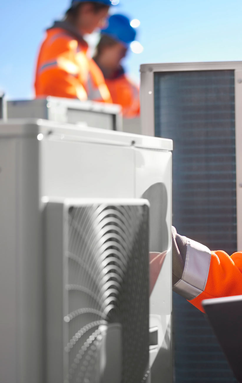 Two workers in orange jackets inspect HVAC units outdoors, with a focus on equipment and safety gear.