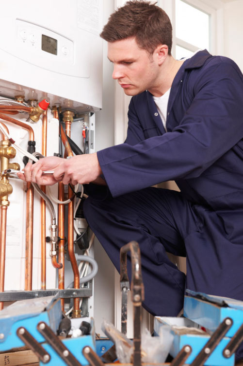 A plumber in blue overalls fixes pipes with tools in front of a boiler in a utility room.