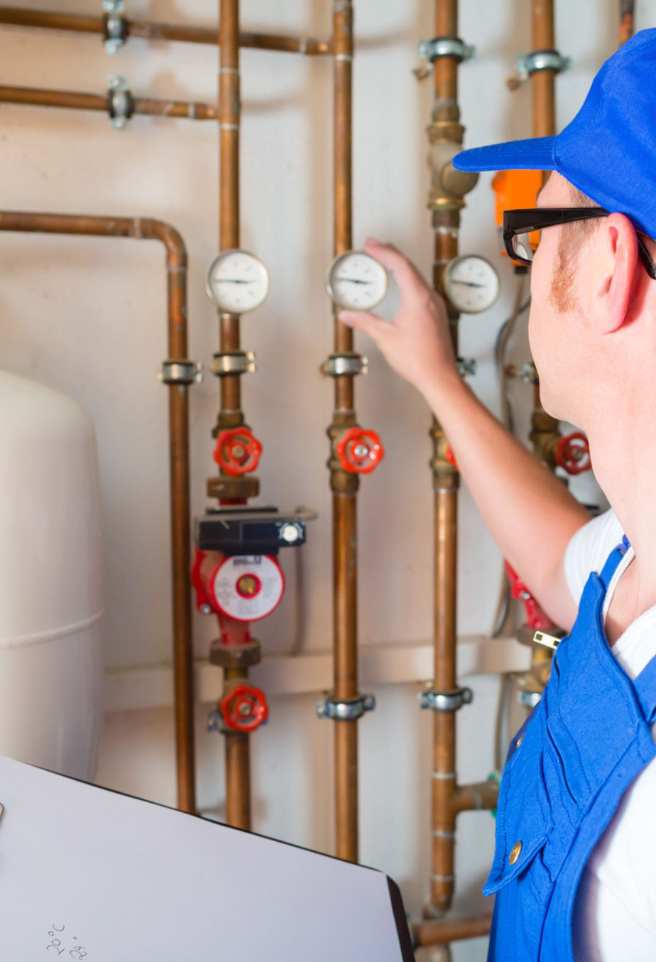 A worker in blue overalls checks pressure gauges on pipes with a clipboard in hand.