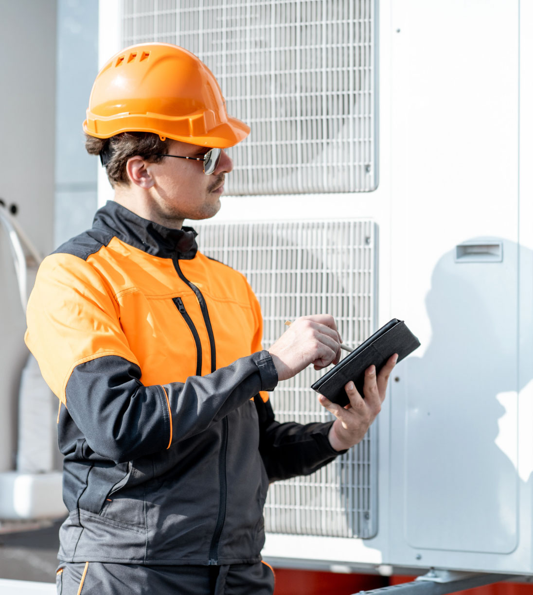 Worker in orange hard hat and jacket uses a tablet while inspecting outdoor industrial equipment.