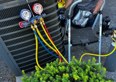 A technician in a gray shirt services an outdoor air conditioning unit connected to manifold gauges.