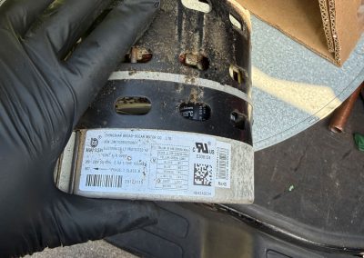 A gloved hand holds a dirty HVAC blower motor with a legible product specification label.