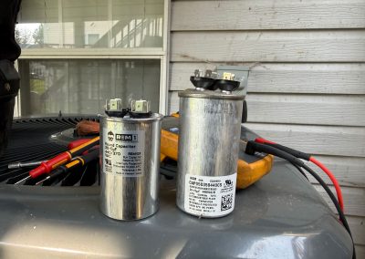 Two cylindrical HVAC run capacitors with electrical terminals, sitting on an outdoor air conditioning unit.