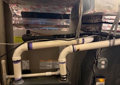 A Goodman HVAC unit with connected metal ductwork and white PVC drainage pipes.